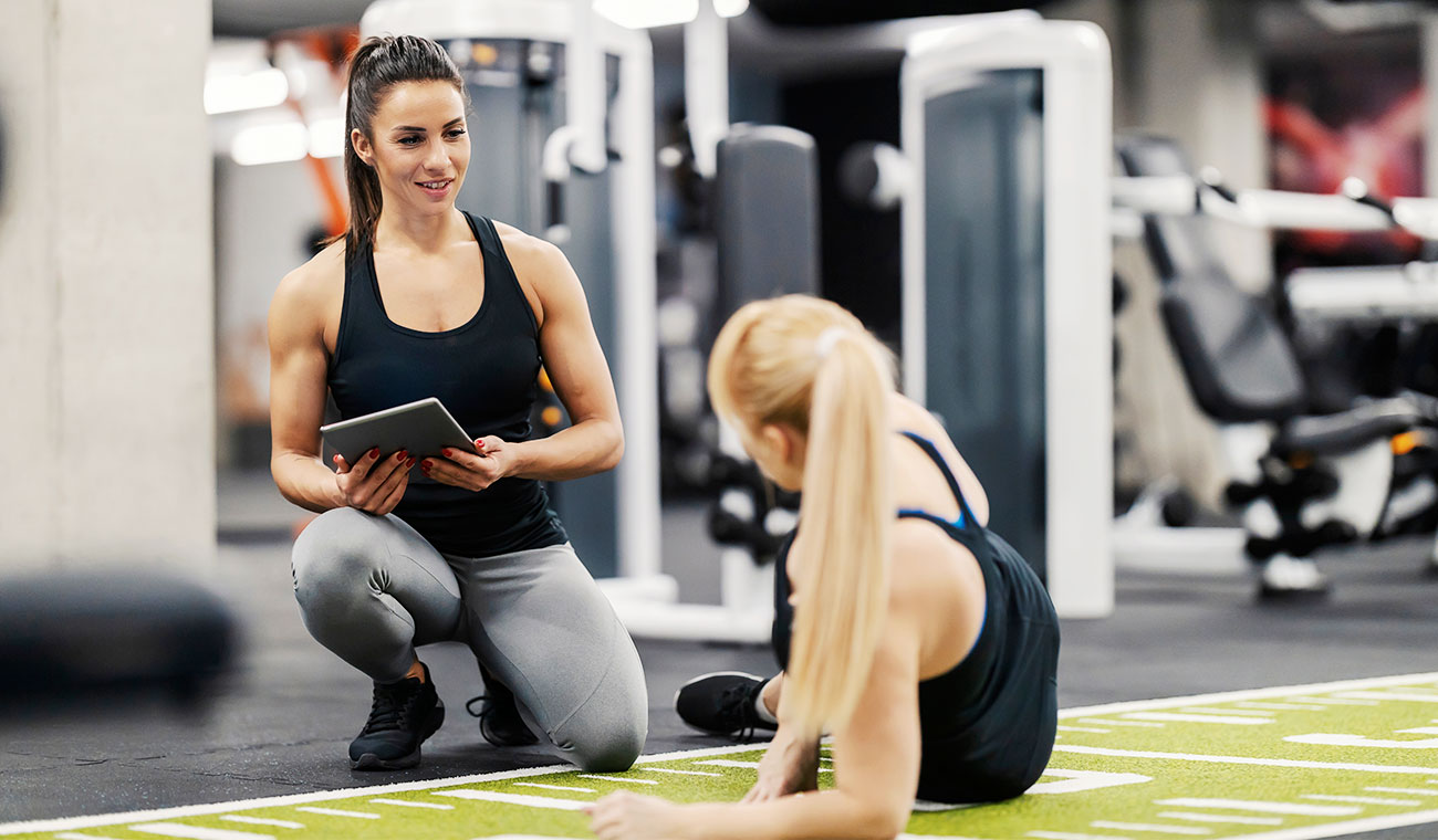 a girl receiving personal training by a muscular trainer in a black tank top holding a tablet