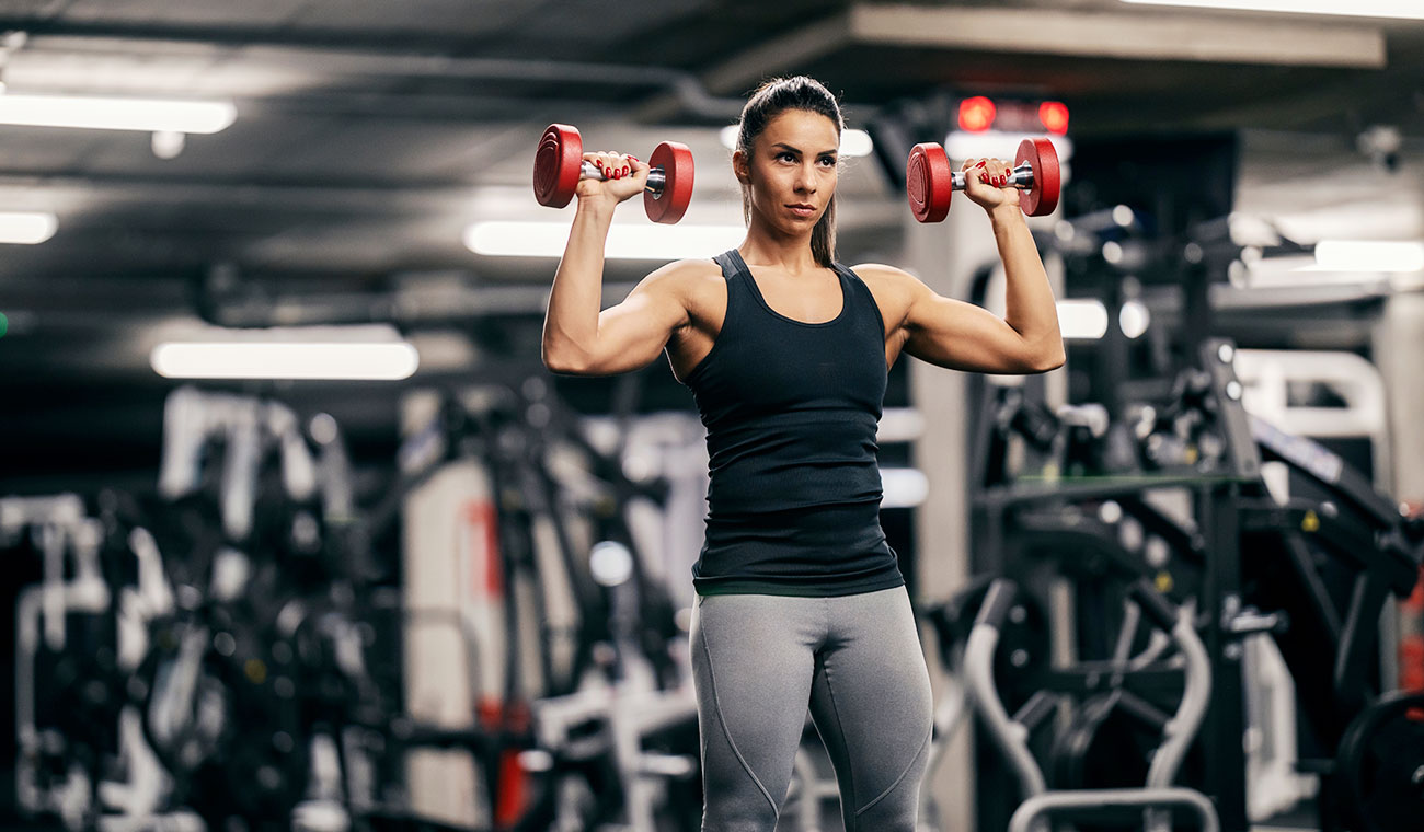 muscular female in a black tank top doing shoulder presses with red dumbells