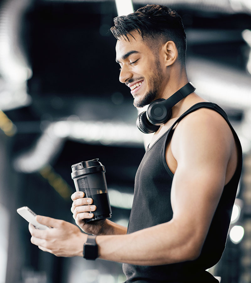 athletic male with headphones around his neck holding a black shaker bottle looking at his phone